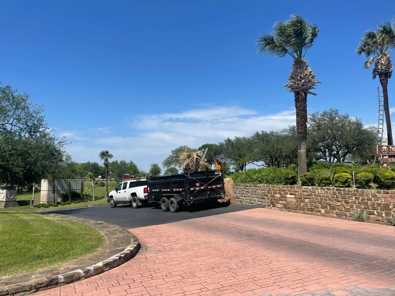 Truck and trailer loaded with palm fronds beside a landscaped entrance with lawn and stone edging